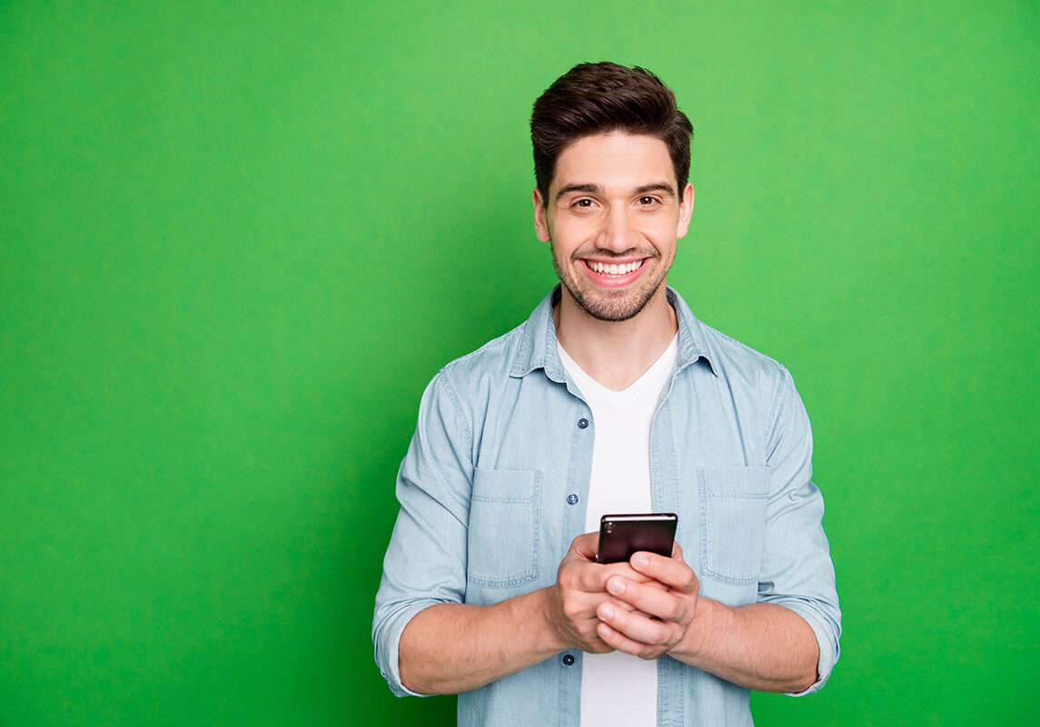 Photo of cheerful brown haired handsome blogger browsing through his telephone, looking for ideas to post in his social media isolated over green vivid color background