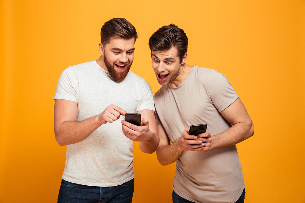 Two joyful men in casual talking and looking at mobile phones in their hands isolated over yellow background